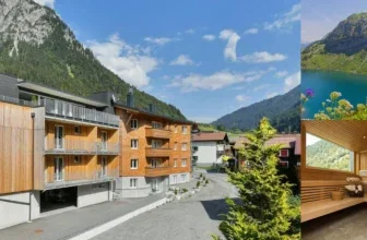 Ein modernes Holzhaus mit mehreren Stockwerken und Balkonen, umgeben von grünen Bergen und einem blauen Himmel.