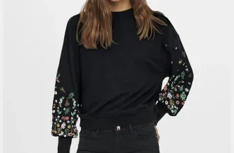Eine junge Frau trägt ein schwarzes Sweatshirt mit Blumenärmeln und schwarze Jeans, vor einem weißen Hintergrund.