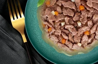 Grünes Tellerchen mit Fleischstücken in Sauce, Karottenwürfeln und Basilikum, daneben goldene Gabel auf schwarzem Stoff