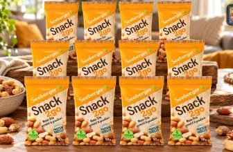 Zwölf orangefarbene Seeberger Snack2go Beutel mit Nuss-Trio-Mix auf einem Holztisch neben einer Schale mit Nüssen.