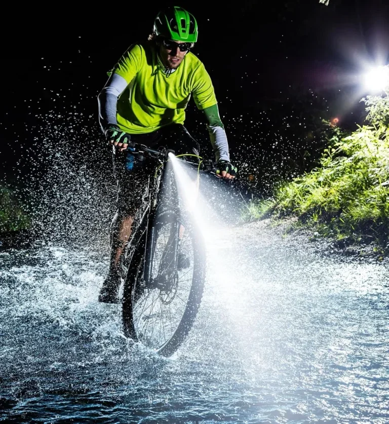 Mountainbiker in Neonkleidung fährt bei Nacht durch Wasser, das Fahrradlicht leuchtet stark.