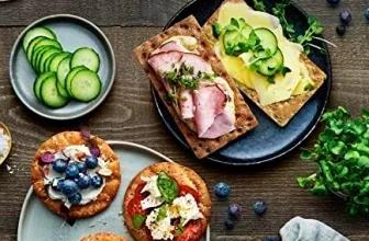 Ein dunkler Holztisch mit verschiedenen Knäckebrot-Variationen, Gurken, Blaubeeren und Kräutern