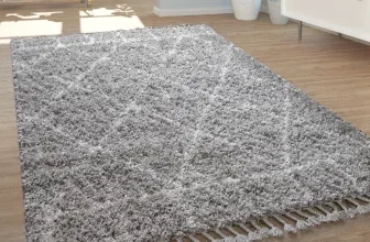 Grauer Hochflor-Teppich mit Fransen auf hellem Holzboden in einem hellen Wohnzimmer.