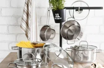 Ein 11-teiliges Topf-Set aus Edelstahl mit Glasdeckeln auf einem Holztisch, im Hintergrund eine weiße Ziegelwand und ein Topfhaken mit aufgehängten Töpfen und einem kleinen Pflanzkübel.