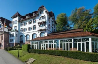 Weißes Hotelgebäude mit braunem Dach und angrenzendem Wintergarten mit Sitzgelegenheiten im Grünen
