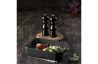 Schwarzes Peugeot Mühlen-Set auf Holzscheit neben Backform mit Gemüse auf dunklem Tisch