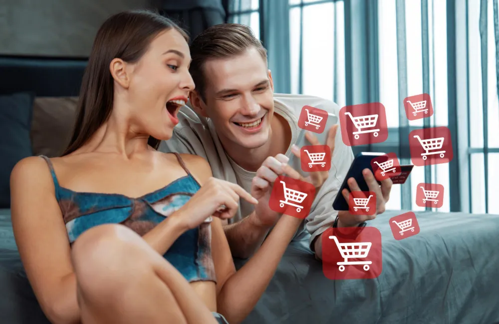 Prime Day Schnaeppchen oder Marketing Trick Junges Paar sitzt auf dem Bett und schaut begeistert auf ein Smartphone, während digitale Einkaufswagen-Symbole um sie herum schweben. Das Bild symbolisiert Online-Shopping, Rabattaktionen und die Jagd nach Schnäppchen während großer Verkaufsaktionen wie dem Amazon Prime Day.