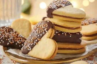 Ein goldenes Tablett mit herzförmigen Sandwichkeksen, die teilweise in dunkle Schokolade getaucht und mit bunten Streuseln bestreut sind.