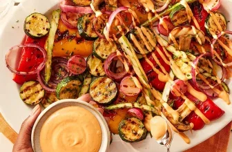 Eine Hand hält eine Schüssel mit einer orangefarbenen Sauce, während die andere Hand einen Löffel in eine Platte mit gegrilltem Gemüse hält.