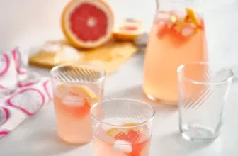 Ein Glas mit einer rosa Flüssigkeit und Eiswürfeln, im Hintergrund ein Krug und eine Grapefruit.