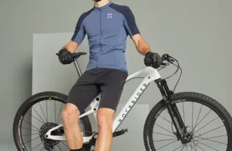 Ein Mann in blauem Radtrikot und schwarzen Shorts sitzt auf einem weißen Mountainbike mit schwarzen Reifen vor grauem Hintergrund