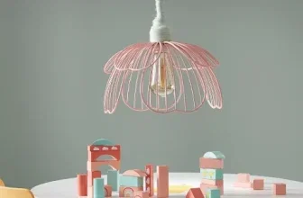 Ein rosa Metall-Lampenschirm in Blütenform hängt über einem weißen Tisch mit Holzbeinen, auf dem farbige Blöcke liegen, daneben stehen ein gelber und ein rosa Stuhl.