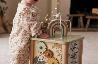 Ein Baby in einem floralen Onesie steht neben einem bunten Holzspielzeug.