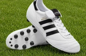 Ein Paar weiße Adidas-Fußballschuhe mit schwarzen Streifen und schwarzen Stollen auf einem grünen Rasen.