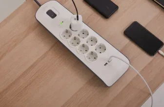 Eine weiße Steckdosenleiste mit 8 Steckdosen und 2 USB-Anschlüssen auf einem Holztisch neben zwei Smartphones und einem schwarzen Gerät.