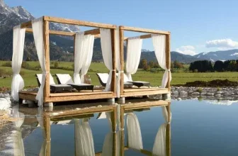 Holzkonstruktion mit weißen Vorhängen und Liegen an einem ruhigen Wasserbecken vor Bergkulisse