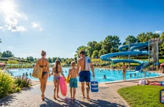 Familie mit zwei Kindern auf dem Weg zu einem Freibad mit großem Schwimmbecken und Wasserrutsche an einem sonnigen Sommertag.