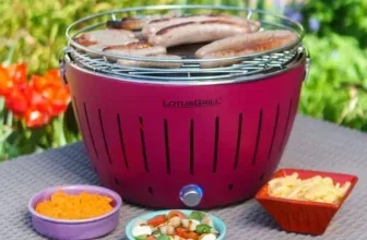 Ein roter LotusGrill Classic mit Würstchen darauf und drei Schüsseln daneben auf einem Tisch im Freien.