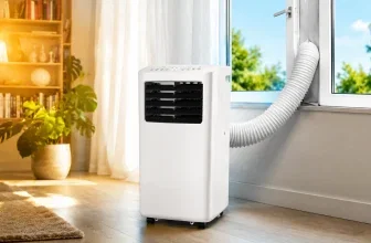 Eine weiße mobile Klimaanlage steht auf einem hellen Boden neben einem Fenster, aus dem ein weißer Schlauch ragt.