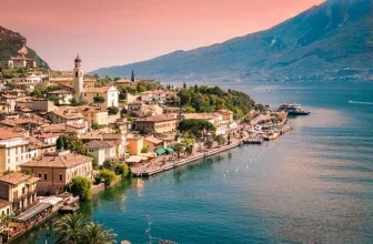 Panoramaansicht eines italienischen Seedorfs am Gardasee mit Hafen, Booten und bewaldeten Hügeln im Hintergrund.