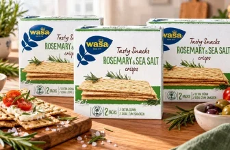 Drei Wasa Rosmarin & Meersalz Crisp-Pakete auf einem Holztisch, daneben belegte Cracker, Tomaten und frische Rosmarinzweige.