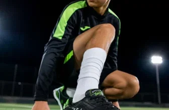 Junge in schwarzer Sportkleidung mit grünen Akzenten hockt auf Kunstrasen und zeigt einen schwarzen Joma Fußballschuh.