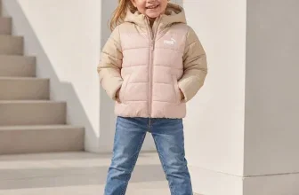 Mädchen in rosa-beiger PUMA Steppjacke, Jeans und weißen Sneakern vor hellem Treppenaufgang
